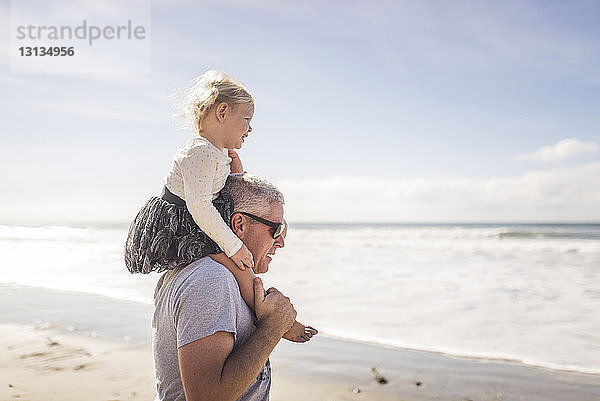 Seitenansicht eines Großvaters  der seine Enkelin auf den Schultern trägt  während er am Strand gegen den Himmel steht