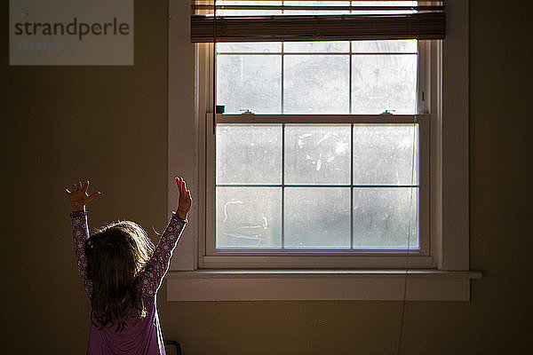 Mädchen mit erhobenen Armen steht zu Hause am Fenster