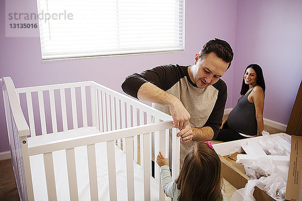 Glückliche schwangere Frau sieht Mann und Tochter an  wie sie zu Hause ein Kinderbett machen