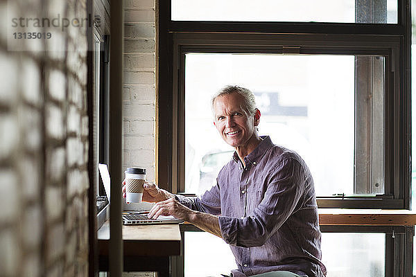 Porträt eines lächelnden reifen Mannes  der eine Kaffeetasse hält  während er seinen Laptop am Tisch im Café benutzt