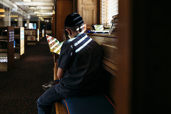 Rückansicht eines Jungen  der ein Buch liest  am Fenster in der Bibliothek