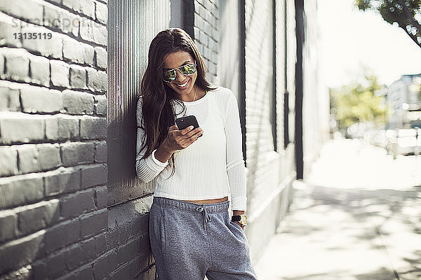 Lächelnde Frau benutzt Telefon  während sie an einem sonnigen Tag an der Wand steht