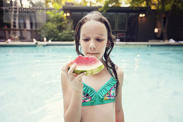 Mädchen isst Wassermelonenscheibe  während sie gegen den Swimmingpool steht