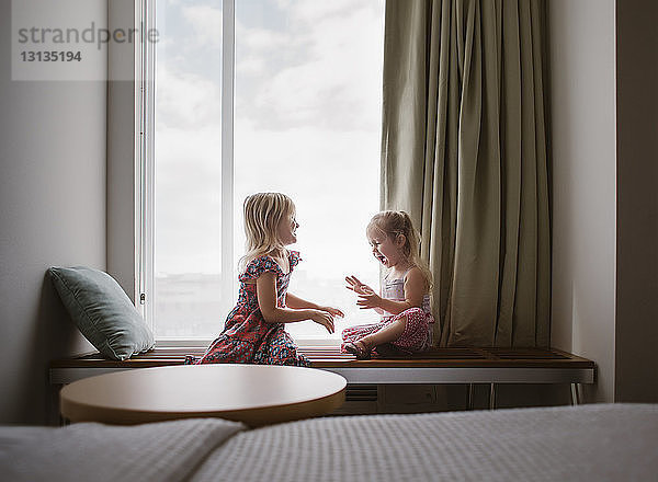 Fröhliche Schwestern reden  während sie zu Hause am Tisch am Fenster sitzen