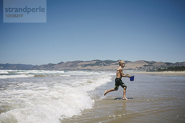Seitenansicht eines Jungen  der Wasser in einem Eimer trägt  während er am Strand an Land läuft
