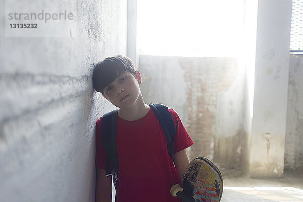 Porträt eines selbstbewussten Studenten mit Rucksack und Skateboard an der Wand in einem alten Gebäude