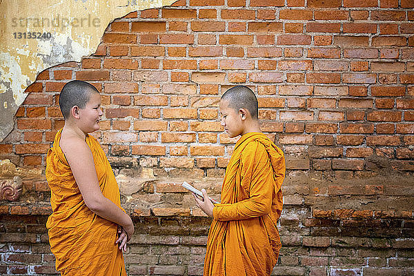 Seitenansicht von buddhistischen Novizenmönchen  die im Stehen an einer Ziegelmauer studieren