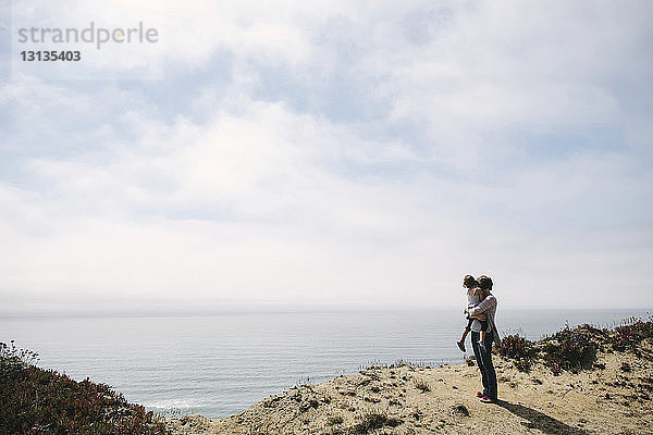 Frau trägt Tochter  während sie auf einer Klippe am Meer gegen den Himmel steht