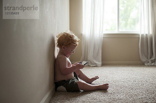 Seitenansicht eines hemdlosen  süßen Jungen  der ein Smartphone benutzt  während er zu Hause auf dem Teppich sitzt