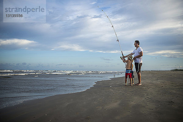 Verspielter Vater und Söhne mit Angelrute stehen am Strand vor wolkigem Himmel