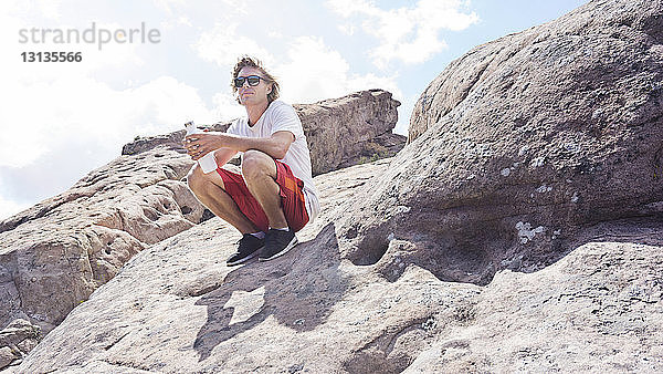 Mann mit Wasserflasche kauert auf einem Felsen vor klarem Himmel