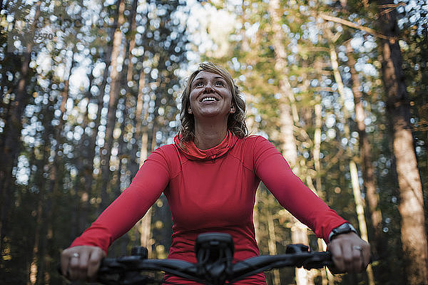 Niedrigwinkelansicht einer fröhlichen Frau beim Mountainbiken gegen Bäume im Wald