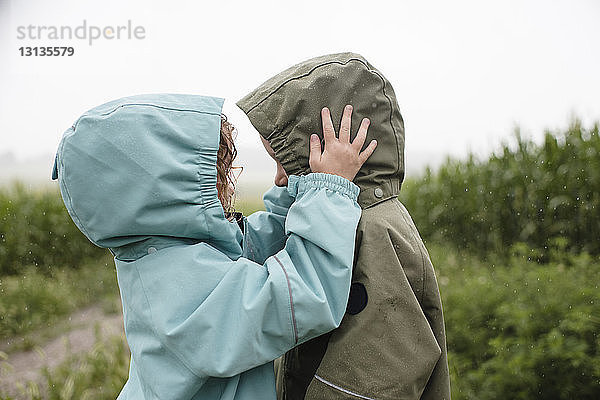 Seitenansicht von Geschwistern in Regenmänteln  die sich gegenseitig ansehen  während sie in der Regenzeit vor Pflanzen stehen