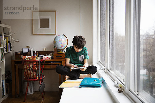 Junge benutzt digitales Tablett  während er zu Hause am Tisch am Fenster sitzt