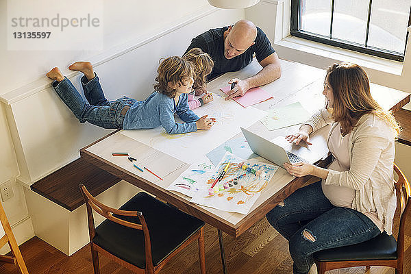 Hochwinkelansicht eines Vaters  der Kindern beim Zeichnen hilft  während die Frau am Tisch einen Laptop benutzt