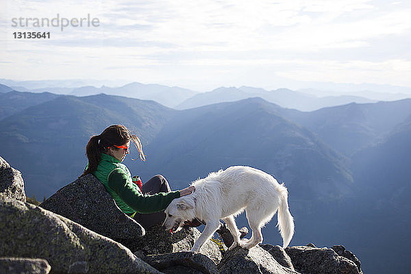 Wanderer streichelt Hund beim Entspannen auf Felsen gegen Bergkette