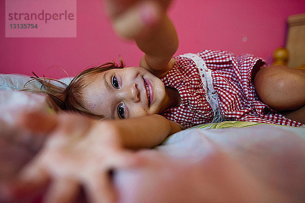 Porträt eines lächelnden Mädchens mit erhobenen Armen  das zu Hause auf dem Bett liegt