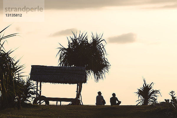 Rückansicht der Silhouette männlicher Freunde  die bei Sonnenuntergang am Strand gegen den Himmel sitzen