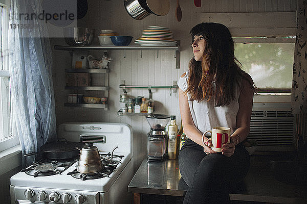 Nachdenkliche Frau hält Becher  während sie auf der Küchentheke sitzt