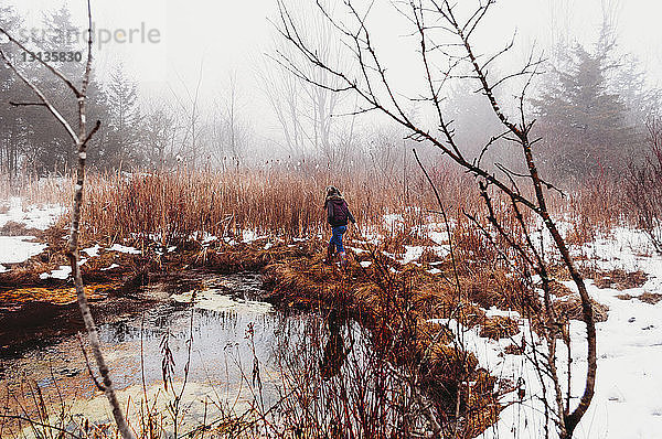 Rückansicht eines Mädchens  das im Winter auf einem Grasfeld am See im Wald spazieren geht