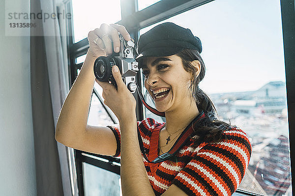 Fröhliche junge Frau fotografiert mit der Kamera  während sie zu Hause vor dem Fenster steht