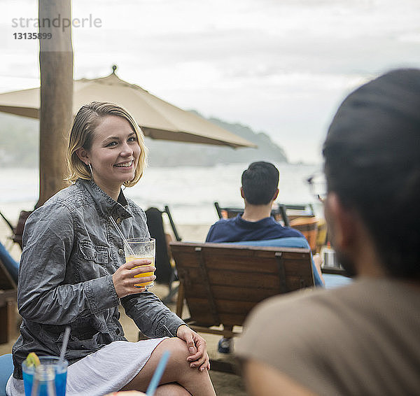 Frau mit Getränk im Gespräch mit einem Freund  während sie am Strand gegen den Himmel sitzt