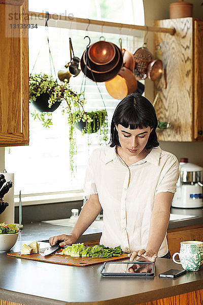 Frau liest digitale Tablette  während sie zu Hause in der Küche frisches Gemüse schneidet
