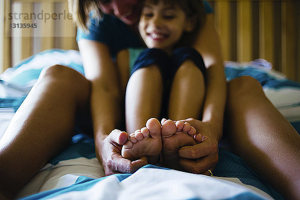 Mutter kitzelt Tochter an den Füßen  während sie zu Hause im Bett sitzt