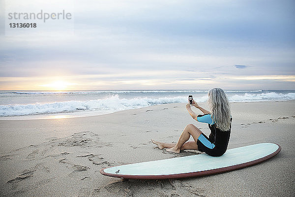 Seitenansicht eines Surfers  der mit einem Smartphone fotografiert  während er auf einem Surfbrett am Strand von Delray sitzt