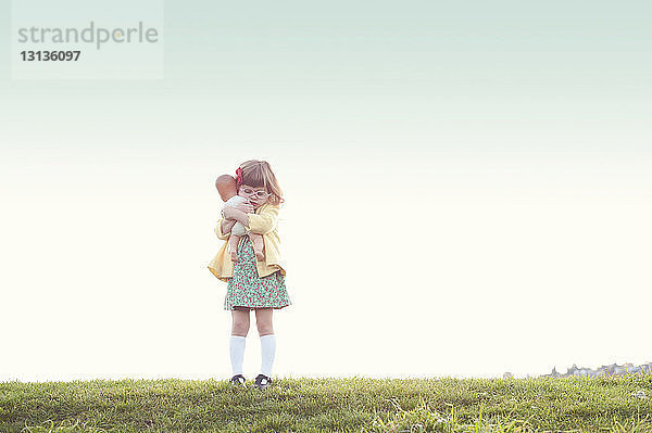 Süßes Mädchen umarmt Puppe  während sie auf dem Feld vor klarem Himmel steht
