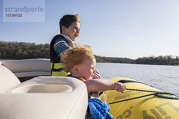 Brüder  die Schwimmwesten tragen  während sie im Boot auf dem See vor klarem Himmel am sonnigen Tag stehen