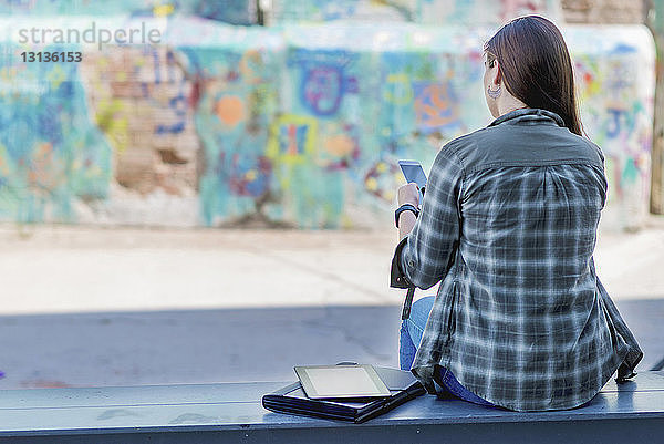 Rückansicht eines Studenten  der ein Smartphone benutzt  während er in der Stadt auf einer Bank sitzt