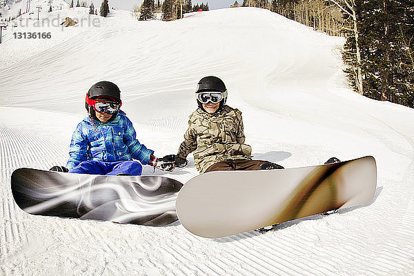 Geschwister tragen Snowboards  während sie auf einem schneebedeckten Feld sitzen