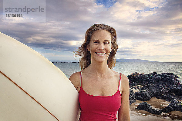 Porträt einer lächelnden Frau mit Surfbrett am Meeresufer vor bewölktem Himmel