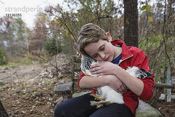 Junge umarmt Huhn  während er auf einem Stuhl im Hinterhof sitzt