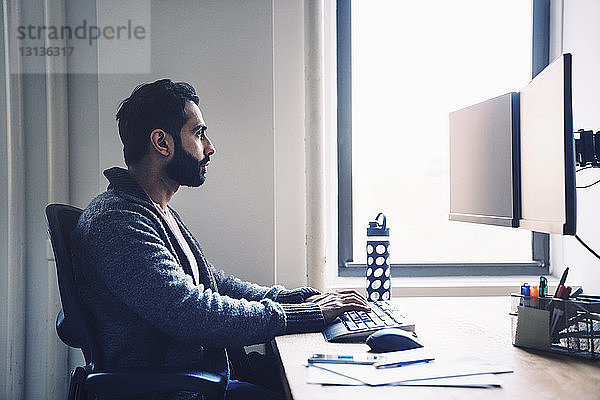 Seitenansicht eines Geschäftsmannes  der einen Desktop-Computer benutzt  während er im Büro am Schreibtisch sitzt