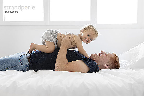 Seitenansicht eines glücklichen Vaters mit hemdloser Tochter  die zu Hause auf dem Bett liegt