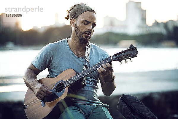 Mann spielt Gitarre  während er bei Sonnenuntergang am Seeufer sitzt
