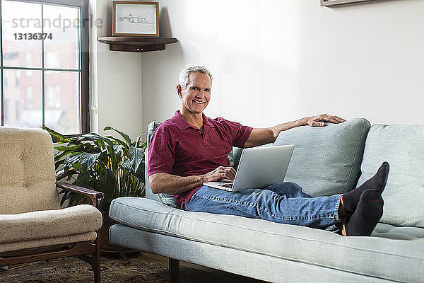 Porträt eines reifen Mannes in voller Länge  der einen Laptop-Computer benutzt  während er zu Hause auf dem Sofa im Wohnzimmer ruht