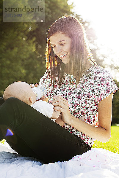 Lächelnde Mutter füttert Baby  während sie im Park sitzt