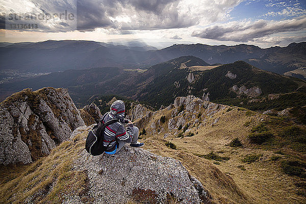 Rückansicht eines männlichen Rucksacktouristen  der auf einem Berg vor bewölktem Himmel sitzt