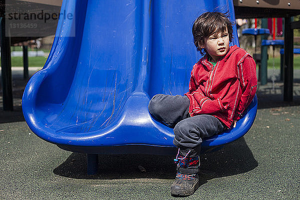 Nachdenklicher Junge schaut weg  während er an einem sonnigen Tag auf einer Rutsche auf dem Spielplatz sitzt