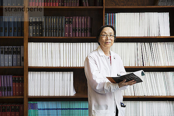 Porträt einer leitenden Wissenschaftlerin  die ein Buch in der Hand hält  während sie im Labor an den Regalen steht