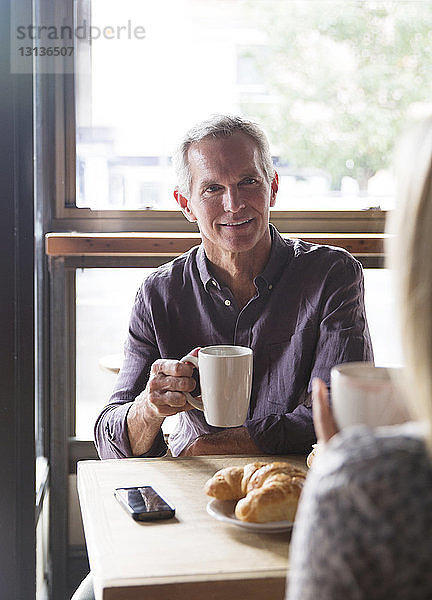 Reifer Mann unterhält sich mit Frau  während er im Café Kaffee gegen das Fenster trinkt
