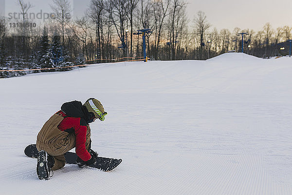 Teenager in voller Länge schnallt bei Sonnenuntergang Snowboard auf schneebedeckter Landschaft