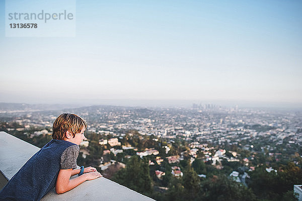 Hochwinkelansicht eines Jungen  der das Stadtbild betrachtet  während er sich auf einer Stützmauer gegen den Himmel auf einer Terrasse lehnt