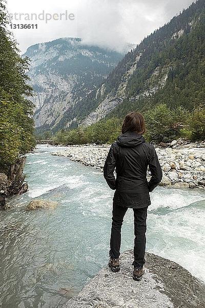 Rückansicht einer Wanderin  die auf Felsen stehend auf Berge und Fluss schaut