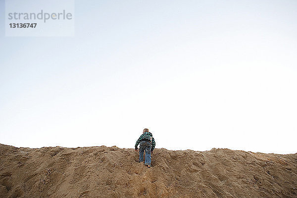 Rückansicht eines Jungen  der auf einer Sanddüne am Strand gegen den Himmel klettert