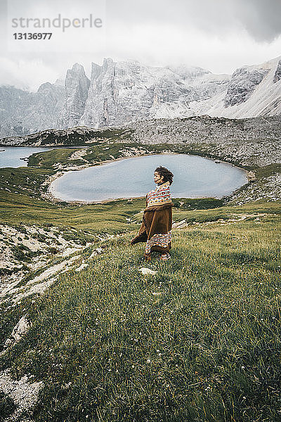Seitenansicht einer Frau mit Decke  die auf einem Grasfeld am See vor bewölktem Himmel steht
