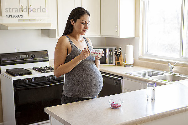 Schwangere Frau frühstückt  während sie in der Küche am Tisch steht
